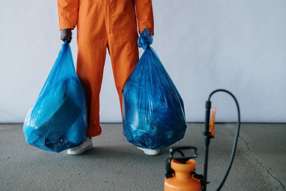 A person dressed in orange protective clothing holding two large blue trash bags filled with waste, standing on a smooth, gray concrete floor next to a wall. In the foreground, a small portion of a portable sprayer or cleaning device with a hose is visible, indicating a cleaning or waste removal process. The scene suggests a professional waste collection or post-cleaning activity, with bright, even lighting highlighting the clean, uncluttered environment. The image emphasizes efficient debris handling as part of domestic or commercial cleaning services provided by Cleaners W10, consistent with surface cleaning and sanitation practices associated with Brompton bulky waste removal and post-clean options in W10.