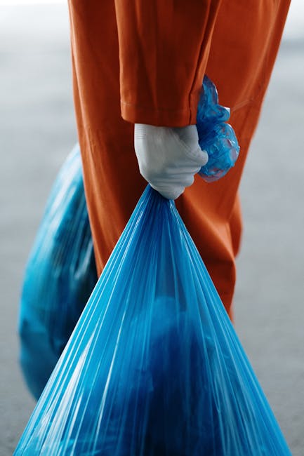 Close-up of a person in orange protective overalls and white gloves holding a tied blue plastic waste bag. The bag is filled with bulky refuse, and the person appears to be engaged in waste collection or removal. The background is blurred, suggesting an outdoor setting, with natural lighting highlighting the clean and organized appearance. This scene exemplifies waste handling procedures, which can be part of comprehensive cleaning and rubbish removal services offered by Cleaners W10, especially relevant to bulk waste removal and post-cleaning disposal in residential or commercial settings.