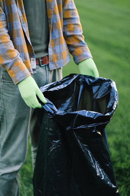 A person dressed in orange protective clothing holding two large blue trash bags filled with waste, standing on a smooth, gray concrete floor next to a wall. In the foreground, a small portion of a portable sprayer or cleaning device with a hose is visible, indicating a cleaning or waste removal process. The scene suggests a professional waste collection or post-cleaning activity, with bright, even lighting highlighting the clean, uncluttered environment. The image emphasizes efficient debris handling as part of domestic or commercial cleaning services provided by Cleaners W10, consistent with surface cleaning and sanitation practices associated with Brompton bulky waste removal and post-clean options in W10.