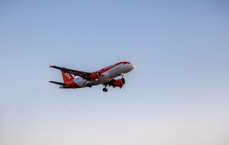 An EasyJet commercial airplane flying in clear blue sky with no visible clouds, captured during ascent or descent. The aircraft has a white fuselage with red accents, including the red winglets and engines, and prominently displays the EasyJet logo on the side. The airplane retains its extended landing gear, indicating it is either approaching or leaving landing or takeoff. Natural sunlight illuminates the plane, highlighting its sleek, aerodynamic shape and the smooth surface of the fuselage. The scene emphasizes the aircraft's clean, well-maintained surface, emphasizing the importance of surface cleaning and maintenance in aviation hygiene, aligning with professional cleaning standards practiced by companies like Cleaners W10.