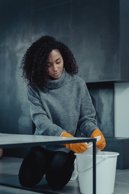 A woman with curly black hair is kneeling on the floor, wearing a grey sweater and orange gloves, and is in the process of cleaning a glass table surface. The room has a modern design with dark grey walls and minimalistic decor. There are cleaning tools and a white bucket nearby, suggesting a deep cleaning or surface cleaning activity. The lighting appears soft, highlighting the reflective quality of the table's glass surface, which is free of dust and smudges. This image represents domestic cleaning services, emphasizing thorough sanitisation and surface maintenance, as offered by Cleaners W10 near Brompton Oratory, SW3, W10, showcasing their expertise in home cleaning solutions.