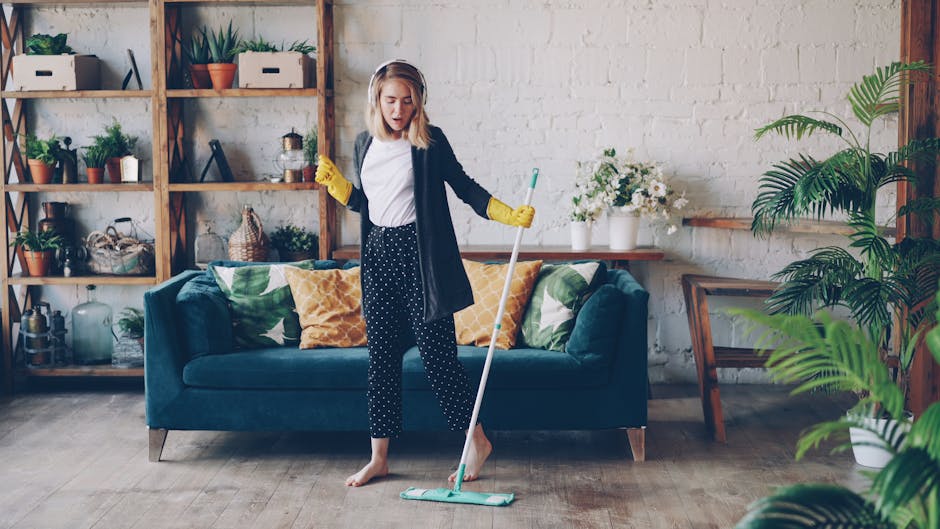 A woman with curly black hair is kneeling on the floor, wearing a grey sweater and orange gloves, and is in the process of cleaning a glass table surface. The room has a modern design with dark grey walls and minimalistic decor. There are cleaning tools and a white bucket nearby, suggesting a deep cleaning or surface cleaning activity. The lighting appears soft, highlighting the reflective quality of the table's glass surface, which is free of dust and smudges. This image represents domestic cleaning services, emphasizing thorough sanitisation and surface maintenance, as offered by Cleaners W10 near Brompton Oratory, SW3, W10, showcasing their expertise in home cleaning solutions.