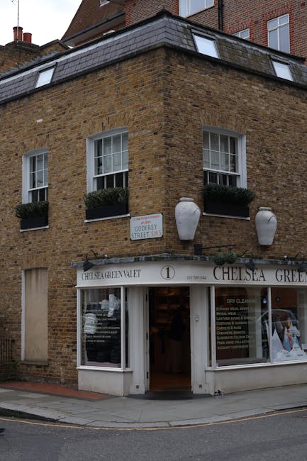 Exterior view of a multi-story brick building housing Chelsea Green Dry Cleaners on the ground floor, with a white sign displaying the shop's name above the entrance. The building features three white-framed sash windows on the upper floor, each adorned with dark green window boxes filled with plants. Two large decorative white ceramic vases are mounted on the brick facade, flanking the signage. The shop's glass front displays cleaning services and promotional signage. The scene is illuminated by natural daylight, with the sidewalk and corner of the street visible in the foreground, emphasizing the building's charming, well-maintained appearance consistent with a clean and inviting commercial property operated by Cleaners W10.