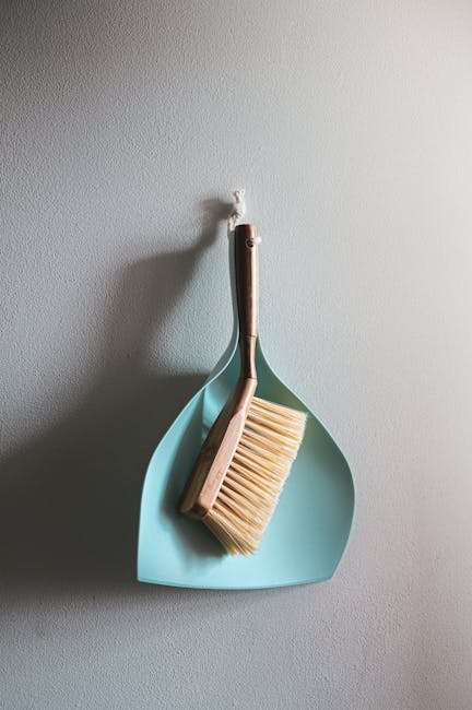 A wall-mounted cleaning tool holder featuring a light blue dustpan and a wooden-handled scrub brush with natural bristles, hanging on a plain white wall. The dustpan has a smooth surface and is clean, with the brush positioned inside it. The setup suggests a tidy, organized space for surface cleaning tools, indicative of domestic or commercial cleaning practices promoted by Cleaners W10 on the page about pricing in Brompton SW3, W10. The lighting highlights the textures and cleanliness of the tools, emphasizing effective surface cleaning and maintenance in a residential or commercial setting.