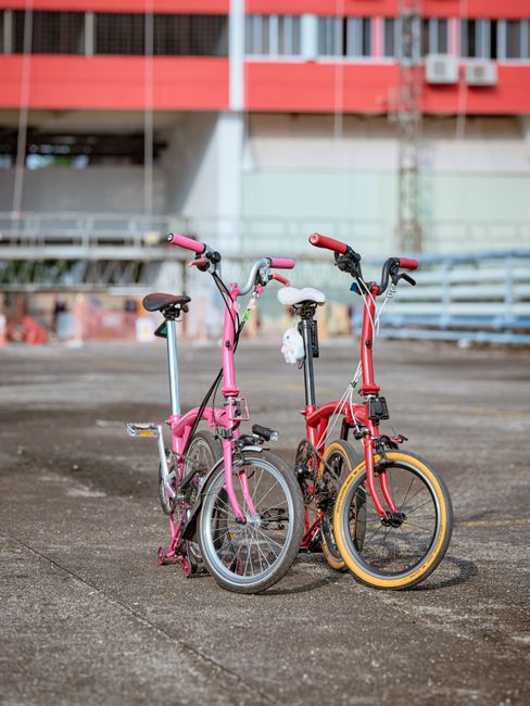 Two children's bicycles, one pink and one red, are parked on a concrete surface in an outdoor urban setting. The pink bike features a small white plush toy hanging from the handlebars, while the red bike has yellow tires and a black seat. In the background, there is a multi-story building with red and gray exterior walls, windows, and air conditioning units, along with some metal railings and a partially blurred urban environment. The area appears clean and well-maintained, with soft natural lighting highlighting the shiny surfaces of the bikes, which are positioned side by side in front of a solid wall or barrier. This image exemplifies surface cleaning and maintenance qualities associated with professional domestic or commercial cleaning services, such as those offered by Cleaners W10.