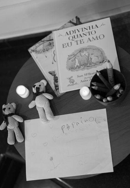 A round wooden table displaying various household items including two small crocheted teddy bears, two white pillar candles, a cup containing coloured pencils, and a children's drawing with the word 'PAPAI MÃO' and small heart doodles. Behind these items are children's books, one with an illustration of a rabbit on the cover titled 'Adivinha Quanto Eu Te Amo' by Sam McBratney. The scene is well-lit, with a clean and organized appearance, emphasizing surfaces like the table and the objects placed on it. Cleaners W10 specializes in surface cleaning and domestic sanitisation, ensuring such spaces remain tidy and hygienic, as illustrated in their service overview on the webpage 'How Much Does an End-of-Tenancy Clean in Brompton Cost?, W10' at cleanersw10.co.uk.