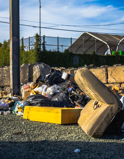 An overflowing waste collection point with a large grey mixed paper and card bin surrounded by numerous black and brown garbage bags, cardboard boxes, and loose waste items scattered on the pavement.