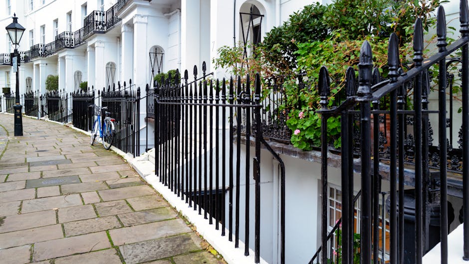 A photograph of a residential street showing a paved sidewalk made of rectangular stone slabs, adjacent to a black wrought iron fence with decorative finials. Behind the fence, there are white terraced houses with black window frames and iron balconies, some with potted plants and flowers. A blue bicycle is parked against the fence. The overall scene appears clean and well-maintained, with the surfaces of the sidewalk and fence free from dirt or debris, and the greenery well-kept. The image emphasizes surface cleaning and maintenance, reflecting the standards discussed in the Landlord Cleaning Standards for Brompton Rentals SW3, W10, as promoted by Cleaners W10.