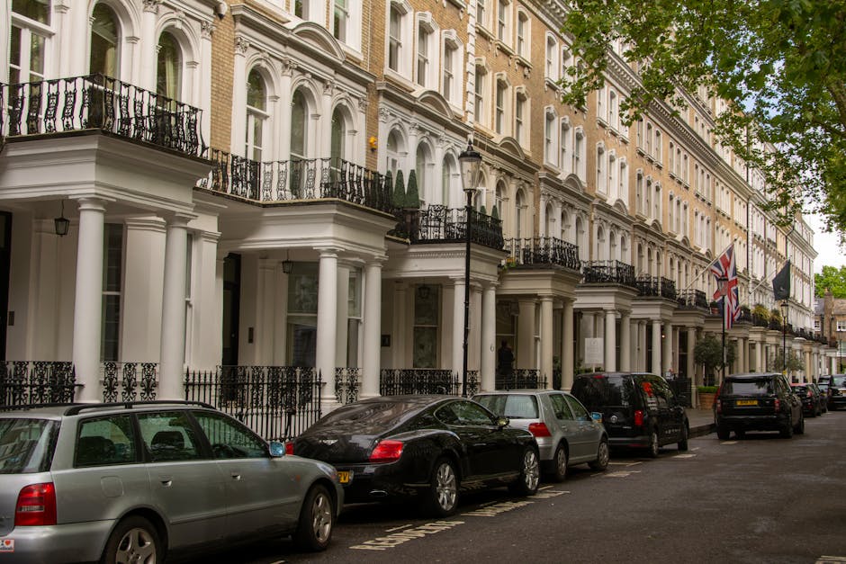A row of elegant Georgian-style terraced houses with white stucco facades, black wrought iron balconies, and tall sash windows on a tree-lined street in Brompton, SW3. The street features parked cars including a silver station wagon, a sleek black coupe, and several other vehicles along the curb. British flags and lampposts line the sidewalk, with lush green trees partially shading the scene. The image captures a bright, clear day, showcasing the well-maintained buildings and orderly street scene, emphasizing a classic residential setting in W10, where professional cleaning by companies like Cleaners W10 ensures the upkeep of such properties.