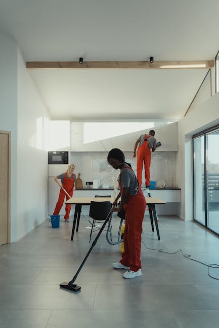 A team of professional cleaners from Cleaners W10 performing surface cleaning in a modern, minimalist kitchen with white walls, a light-colored tiled floor, and large floor-to-ceiling glass sliding doors. The cleaners, dressed in red uniforms, are using various tools: one individual is vacuuming the floor with a handheld vacuum, another is wiping the kitchen countertop with a cloth, and a third is cleaning a high cabinet or ceiling area using a step ladder. The natural light floods the space through the glass doors, highlighting the clean, polished surfaces and the orderly arrangement of the room's sleek cabinets and appliances. This scene exemplifies thorough domestic cleaning and surface sanitisation as part of a same-day emergency cleaning service in Brompton, provided by Cleaners W10.