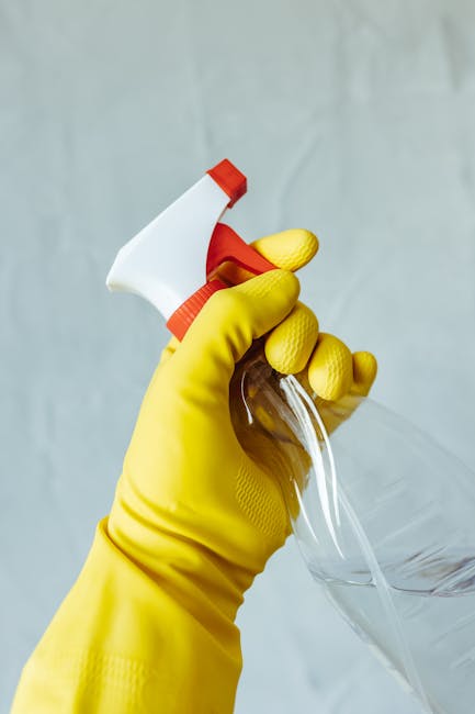 Two professional cleaners from Cleaners W10 standing side by side against a plain white wall in a well-lit room. The cleaner on the left is holding a vacuum cleaner with a black hose, wearing a beige uniform with rolled-up sleeves, and has dark hair pulled back. The cleaner on the right is holding a mop with a green handle, wearing a similar beige uniform, with short, platinum blonde hair, and is smiling. The scene suggests a focus on domestic cleaning services, with a clean and organized appearance suitable for surface cleaning, sanitisation, and deep cleaning tasks in residential or commercial settings.