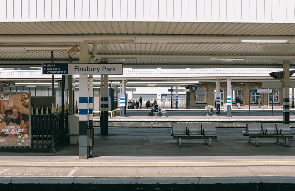 At Finsbury Park station, the platform features polished concrete floors that appear clean and well-maintained, with visible lines marking the edge for safety. The station's ceiling is paneled with white, corrugated material, illuminated by long, fluorescent lighting fixtures. Metal benches with wooden seats are arranged along the platform, showing no litter or debris, indicating regular cleaning. The station signage, including a prominent white sign with black lettering reading 'Finsbury Park,' is visible hanging from the ceiling. Behind the benches, more platforms and tracks extend into the background, with a few passengers seated or walking, all appearing orderly and hygienic. The overall scene reflects thorough surface cleaning and maintenance, aligning with the standards of domestic and commercial sanitisation by Cleaners W10, ensuring a safe and clean environment for travelers.