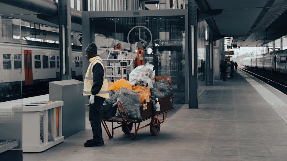 At West Brompton Station in the W10 area, a worker wearing a dark jacket, grey beanie, and high-visibility vest is standing on the platform beside a large cart filled with black, grey, and orange trash bags. The station features smooth, dark grey tiled flooring, modern glass walls, and metal support structures. In the background, trains are visible on either side, with station signage and a few people further along the platform. Natural lighting illuminates the scene, highlighting the clean and orderly appearance of the station. The image reflects a focus on maintaining cleanliness in a busy transportation hub, consistent with professional station cleaning and hygiene standards, as emphasized by Cleaners W10.